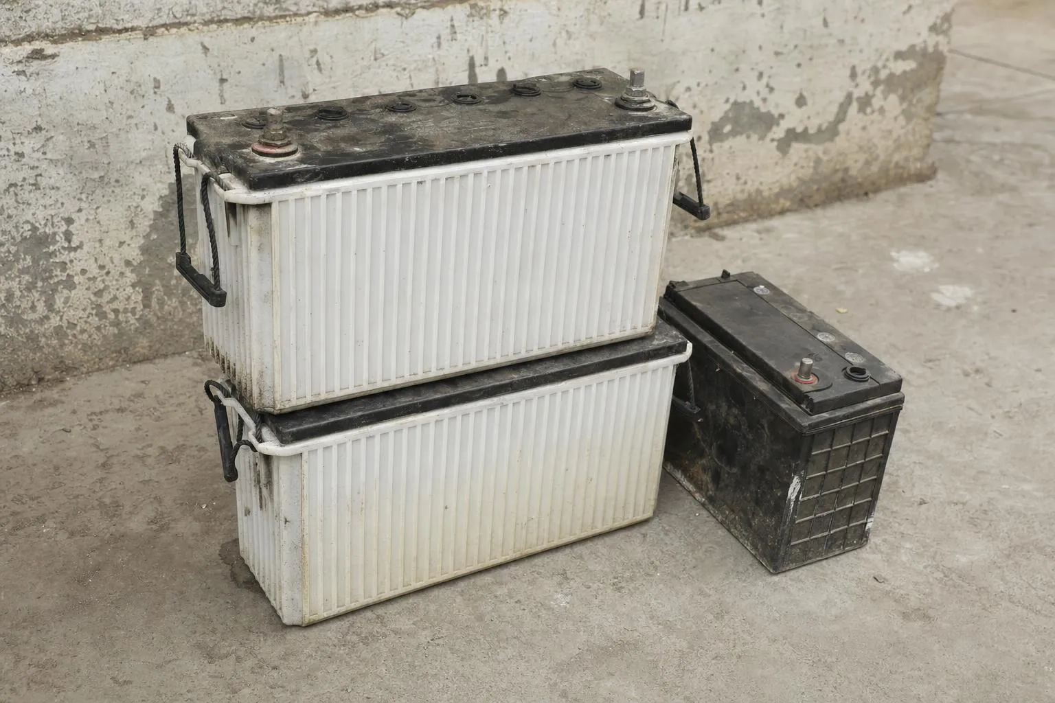 Old lead-acid batteries stacked on a concrete floor: two large inverter batteries with visible terminals beside a smaller car battery, dusty and worn